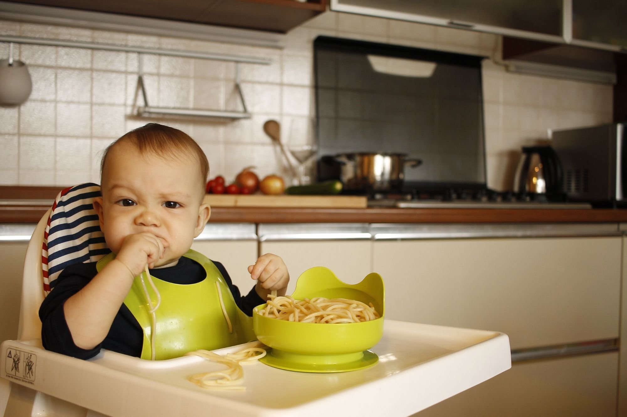 Toddler in a highchair eating pasta from a green bowl