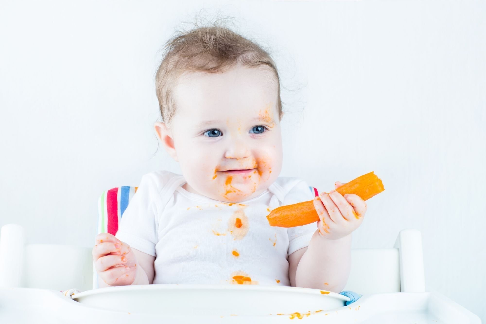 Toddler holding a soft carrot, with a food stained face and vest