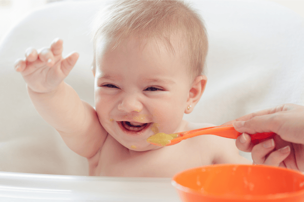 Happy baby raising one hand in the air while his parent is spoon feeding him