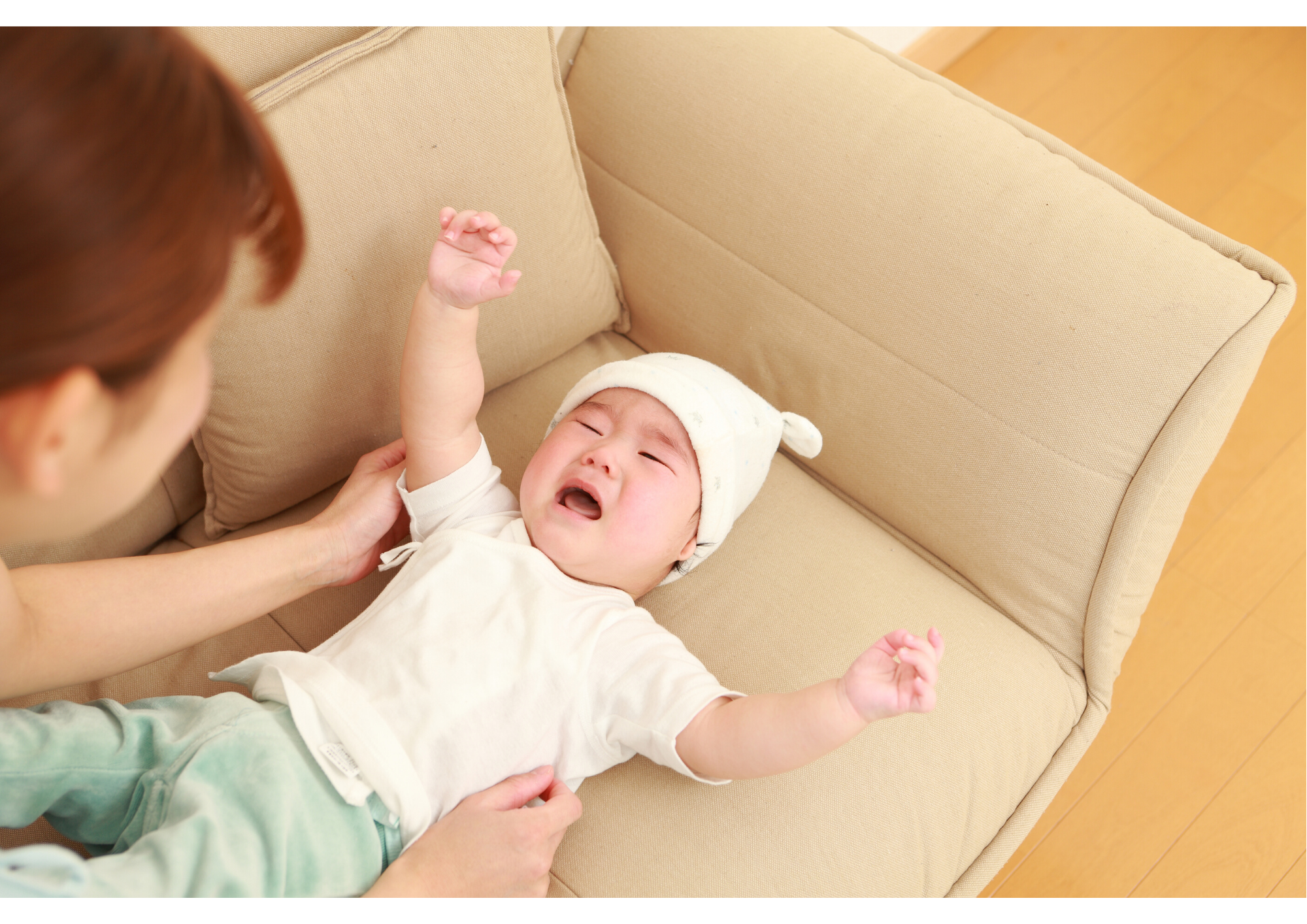 Parent bending over to pick up their crying baby who is laying on the couch holding his arms up