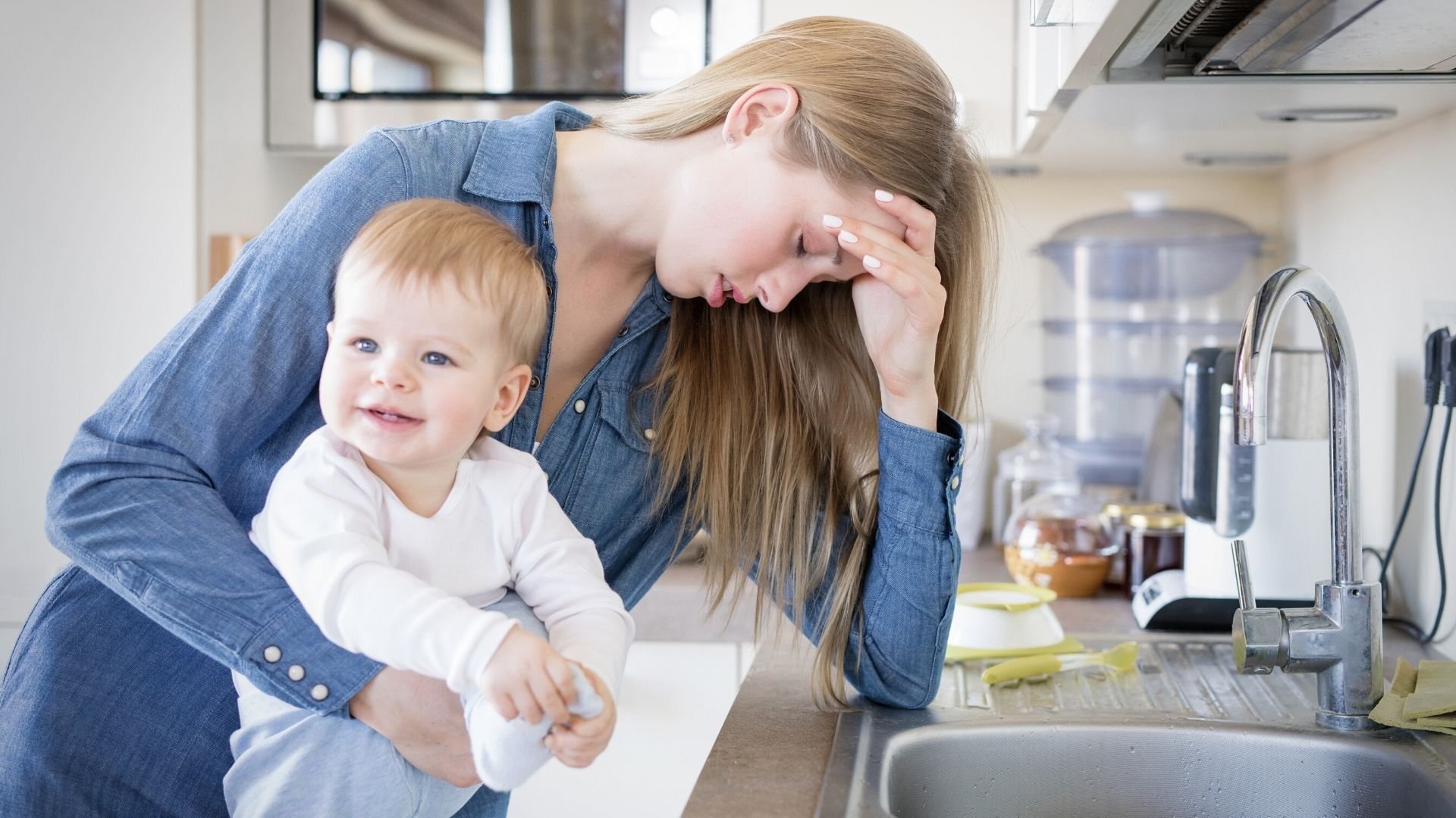 Tired mum leaning on the edge of the sink with her head in her hand, holding her baby in her other arm