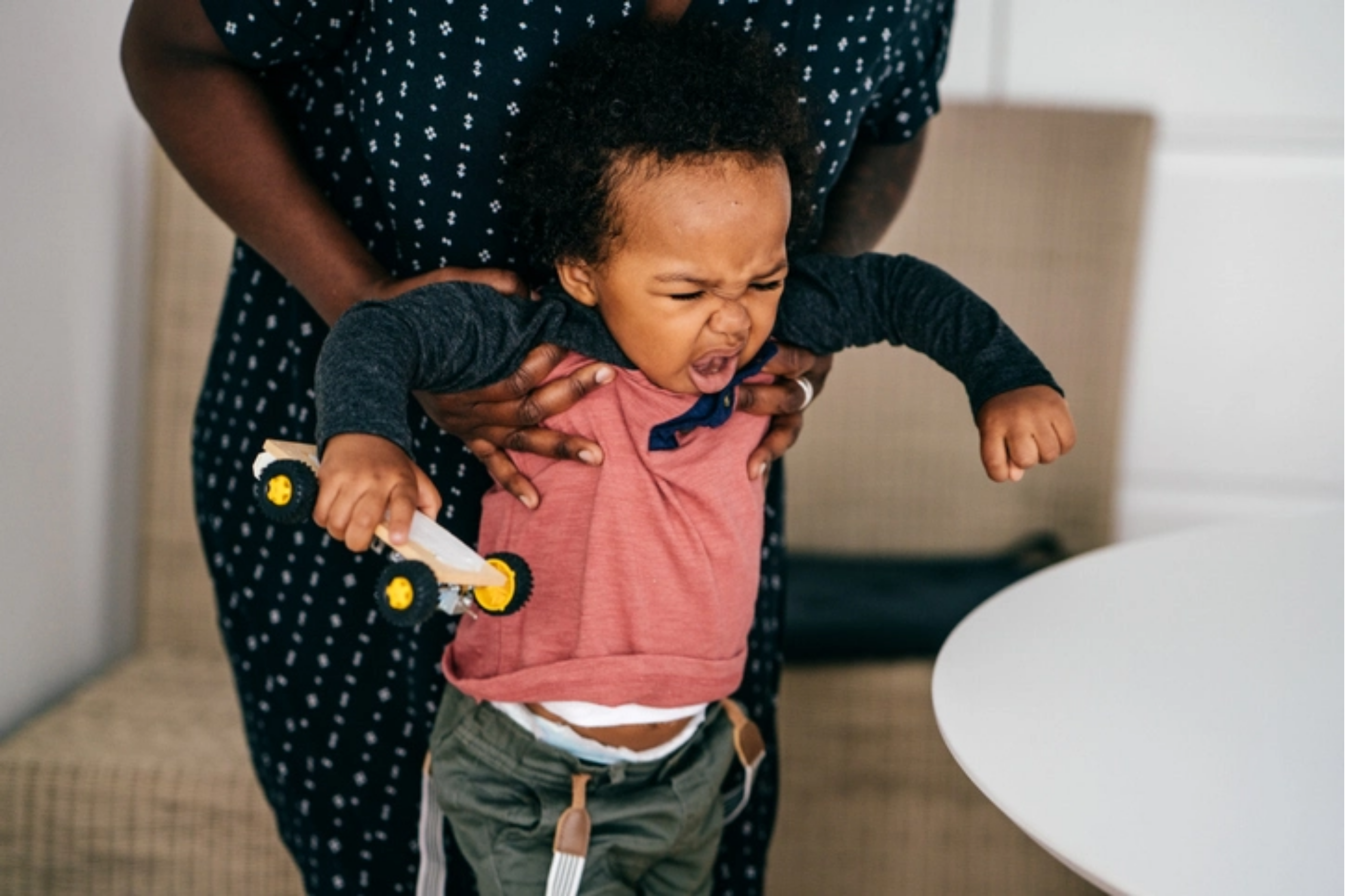 Toddler wearing a pink and navy top throwing a tantrum in the dining room while her parent is holding her under her arms