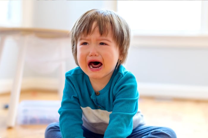 A toddler crying while sitting on the floor, expressing frustration or distress in a brightly lit room.