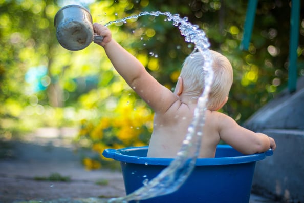 Baby in a blue bathing tub outdoors, splashing water from a mug