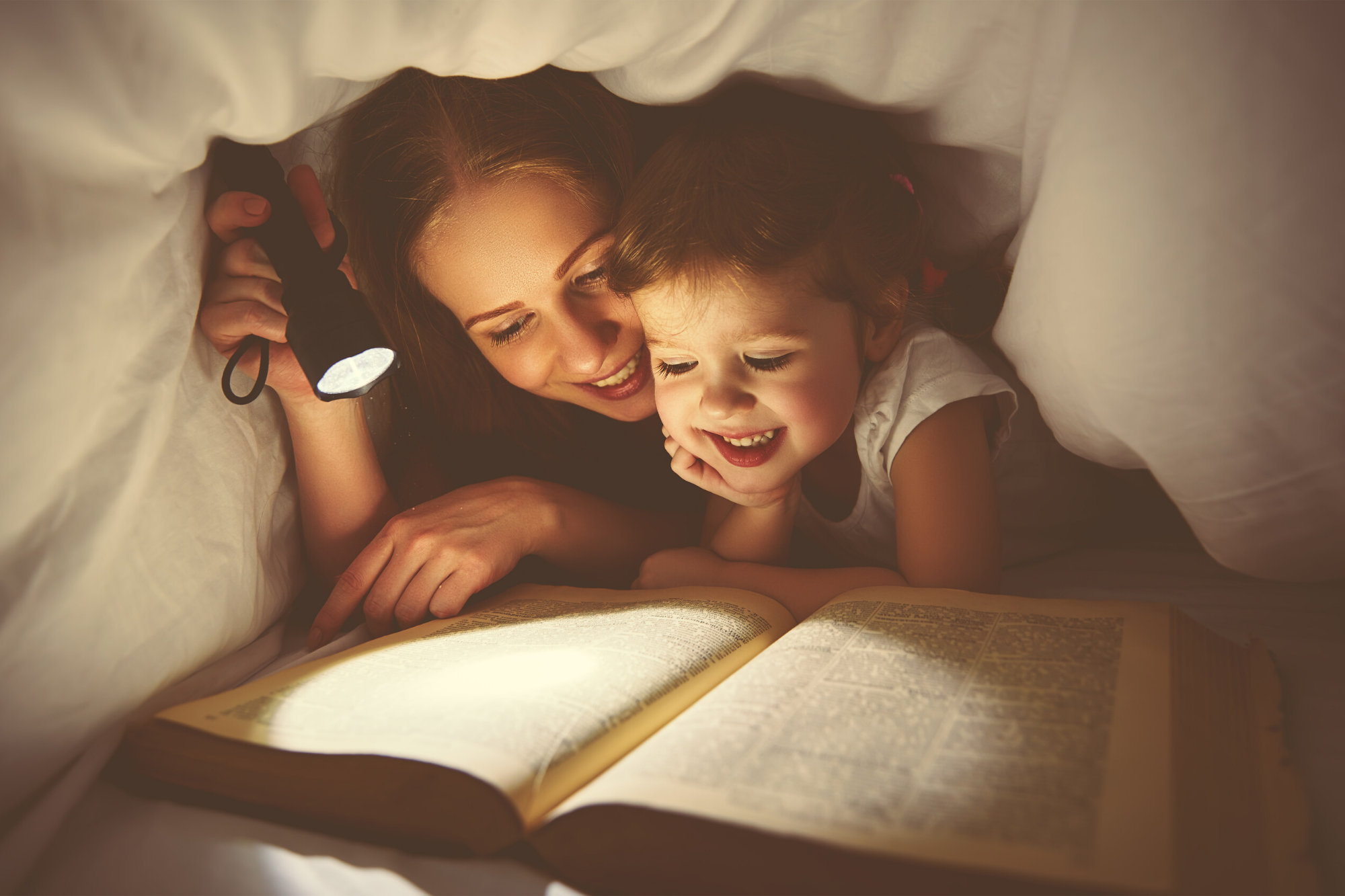 Mum with a torch reading to her child underneath bed covers