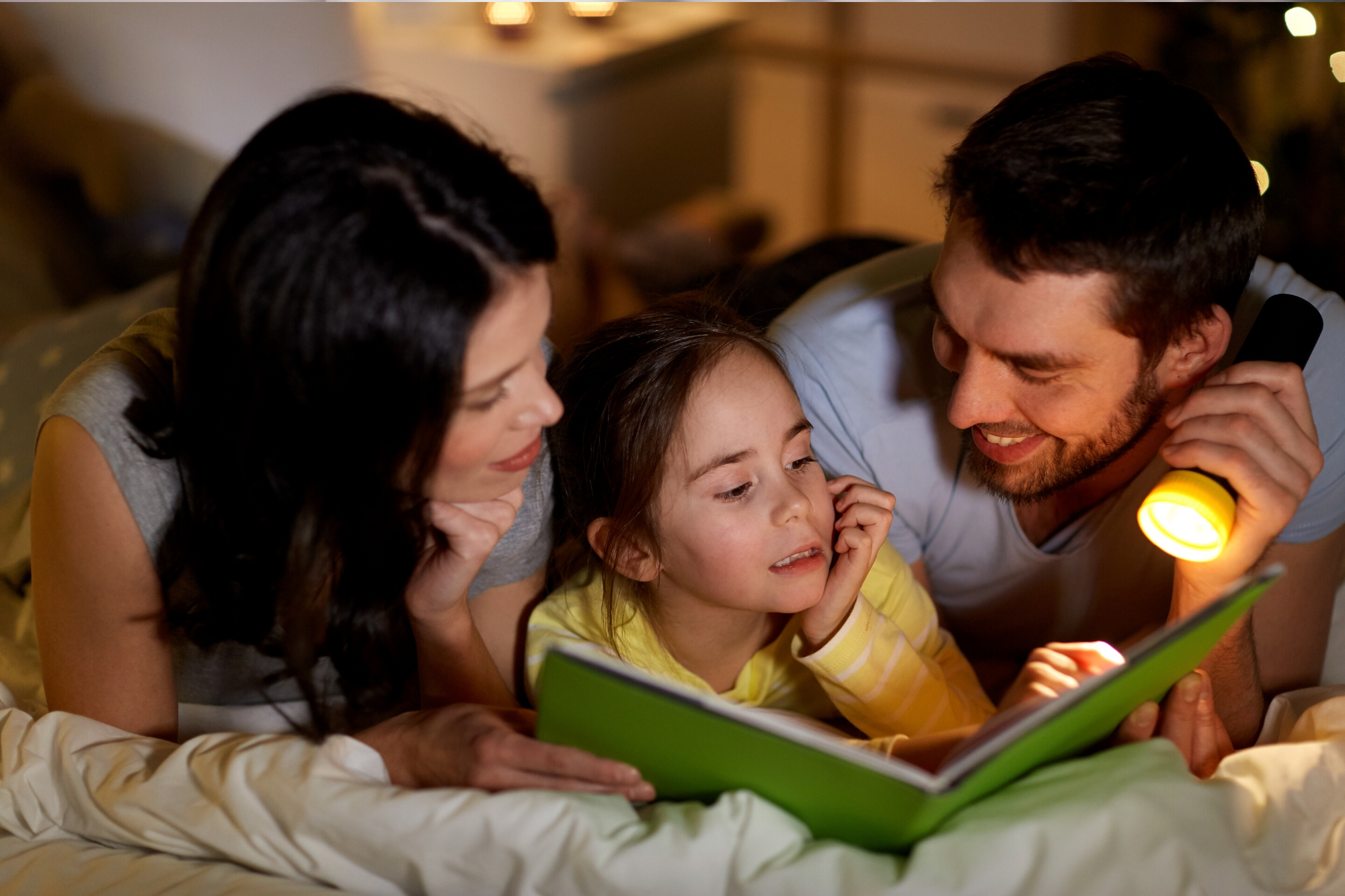 Dad and mum shining a torch on a book in bed with their toddler
