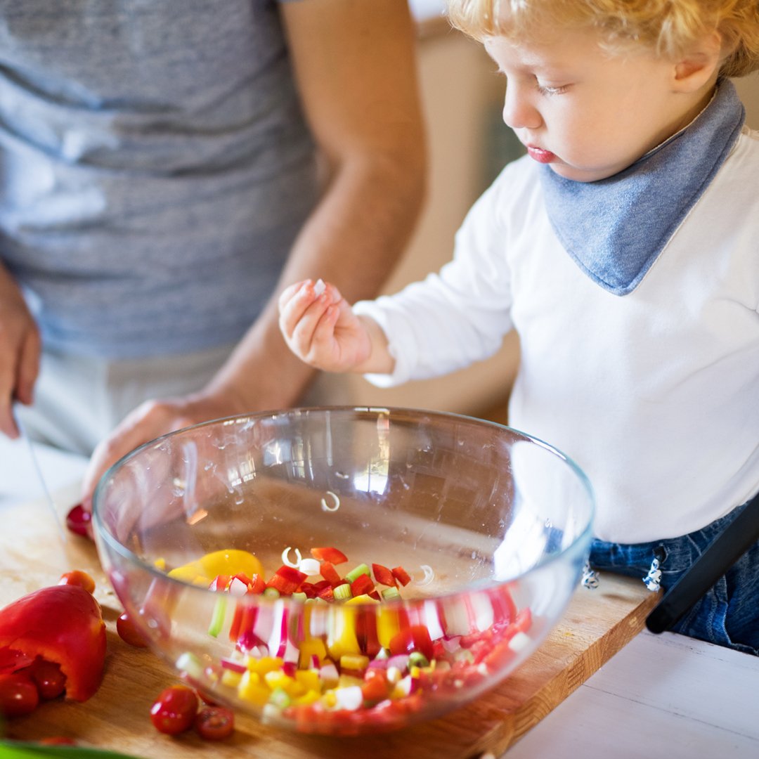 Toddler wearing a white top and blue handkerchief around his neck holding some chopped peppers in the kitchen