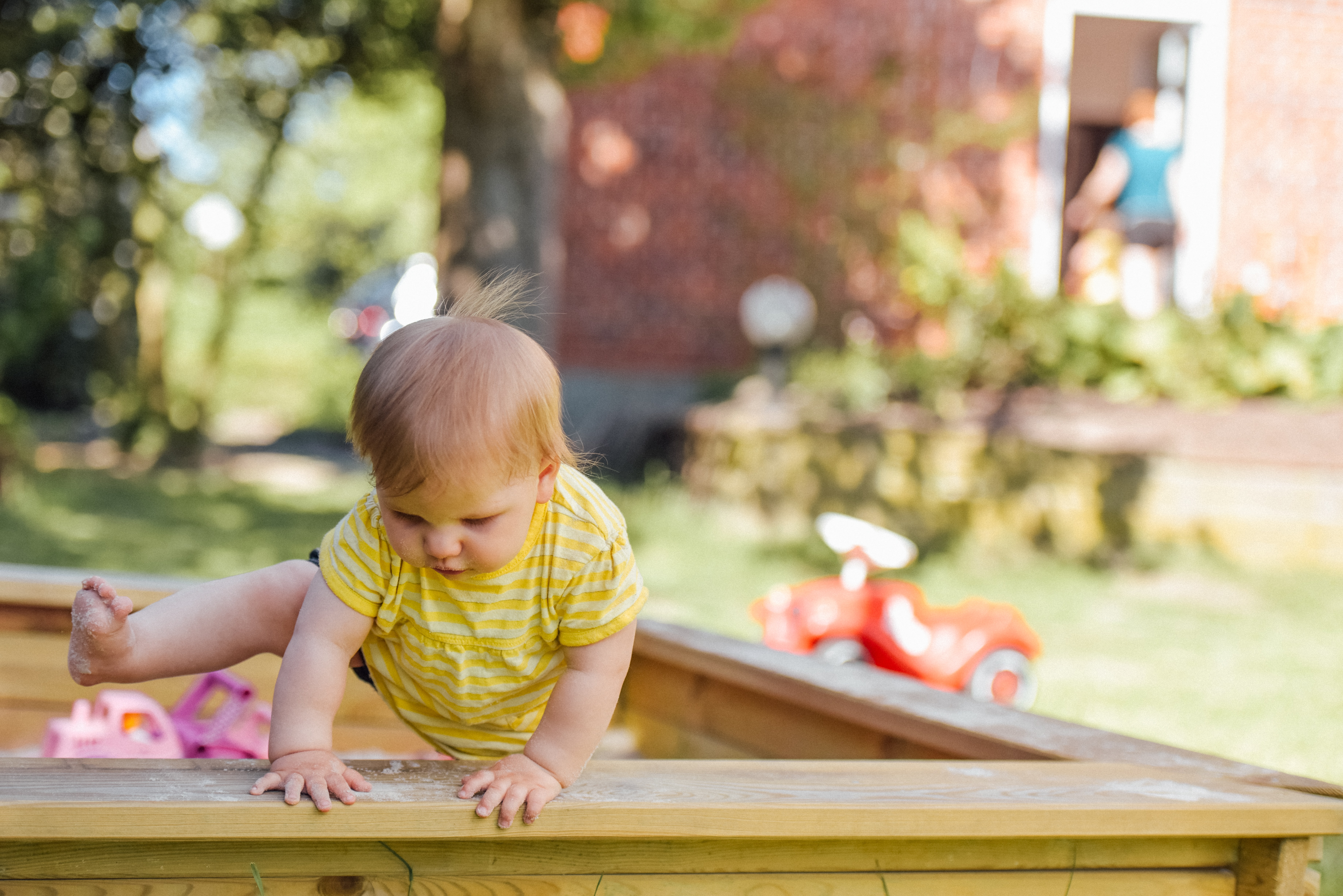 Baby girl wearing a yellow top climbing over a wooden playpen in the garden