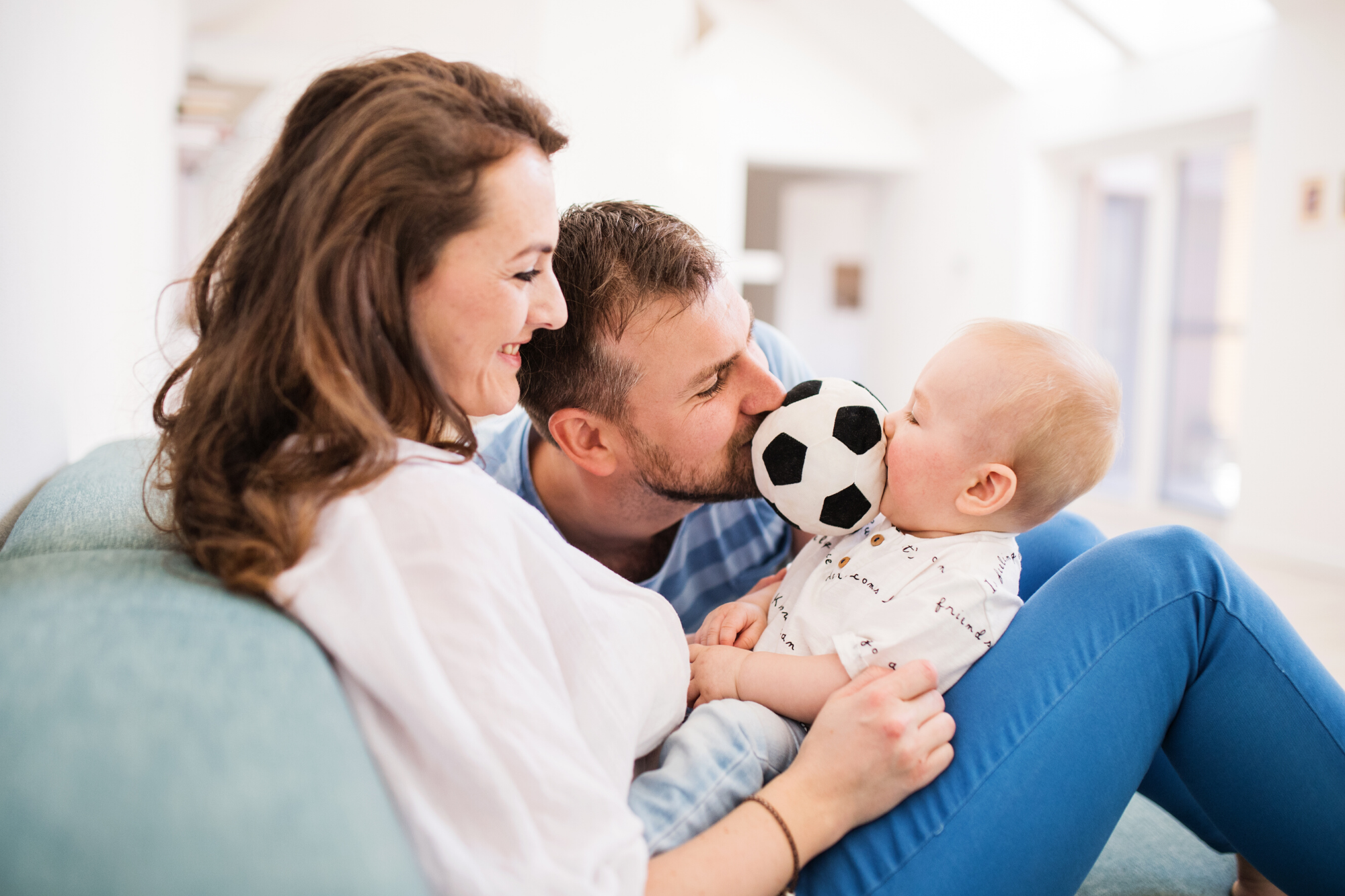 Baby sitting between mums legs, dad is holding a plush football toy between his mouth and the babys mouth