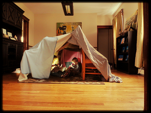 Siblings sitting inside a homemade fort in their living room, reading with a torch