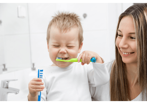 Toddler scrunching his face, while he is brushing his teeth, mum is smiling as she supervises