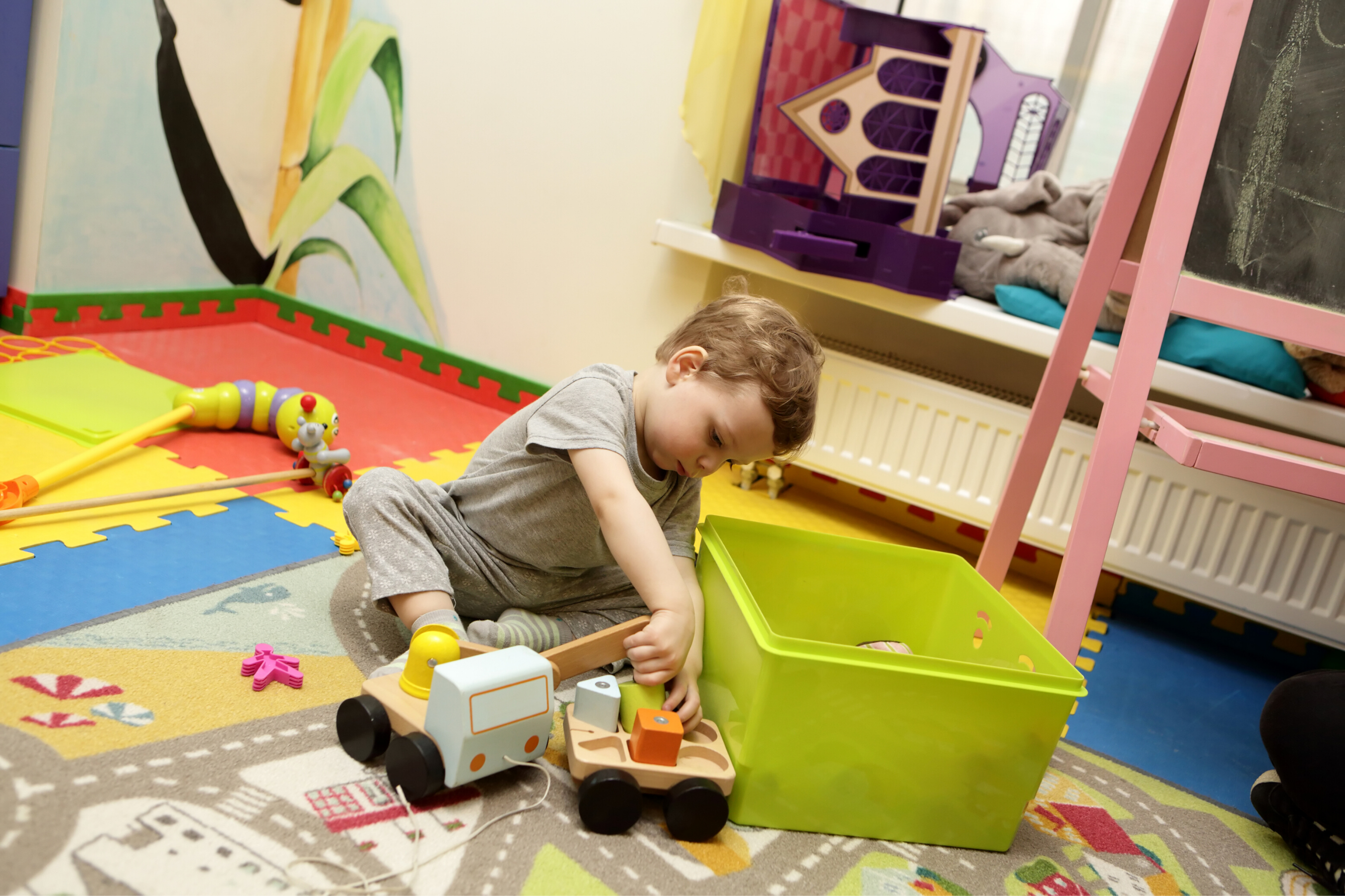 Little boy wearing a grey outfit, playing with toy cars beside a green plastic box