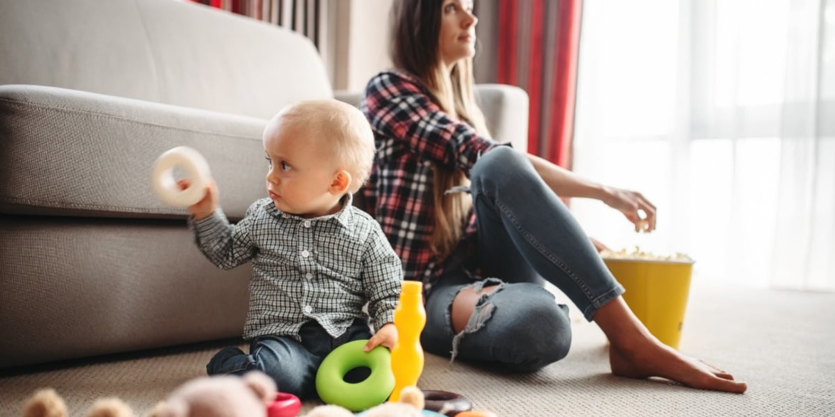 Baby in checked shirt playing with ring toys in the living room while mum takes a break