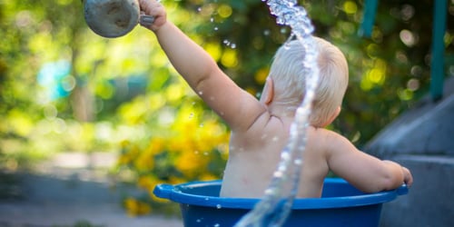 Baby sat in a blue plastic tub holding a watering can filled with water