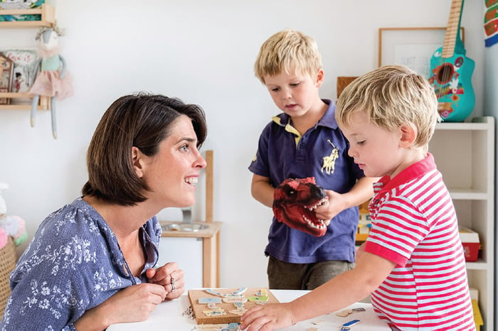 Mother engaging with her two young children at a table, supporting their creativity and play with puzzles and a toy dinosaur in a bright and playful home environment.