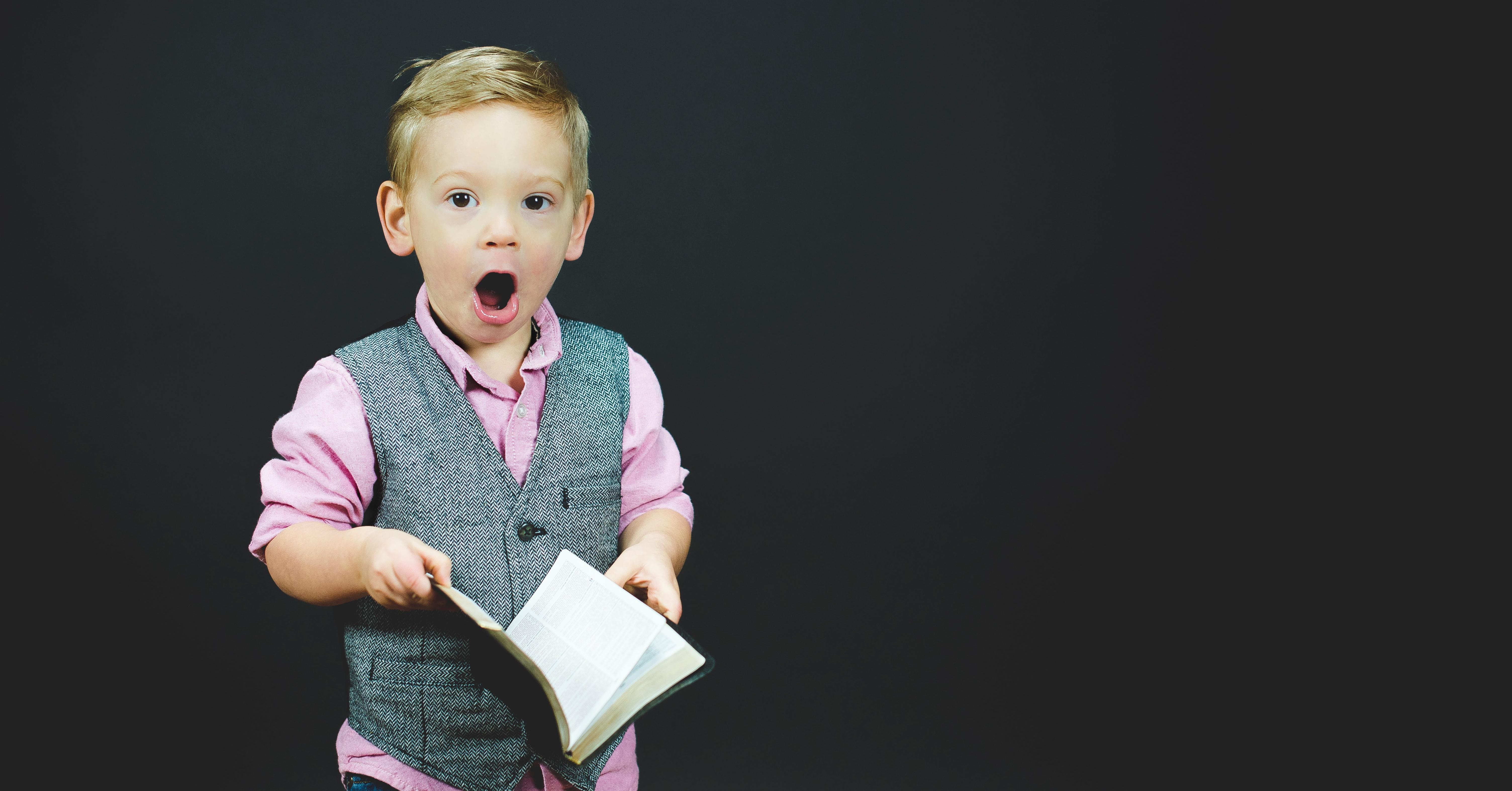 Child standing holding a book, mouth wide open dressed in a waistcoat and pink shirt
