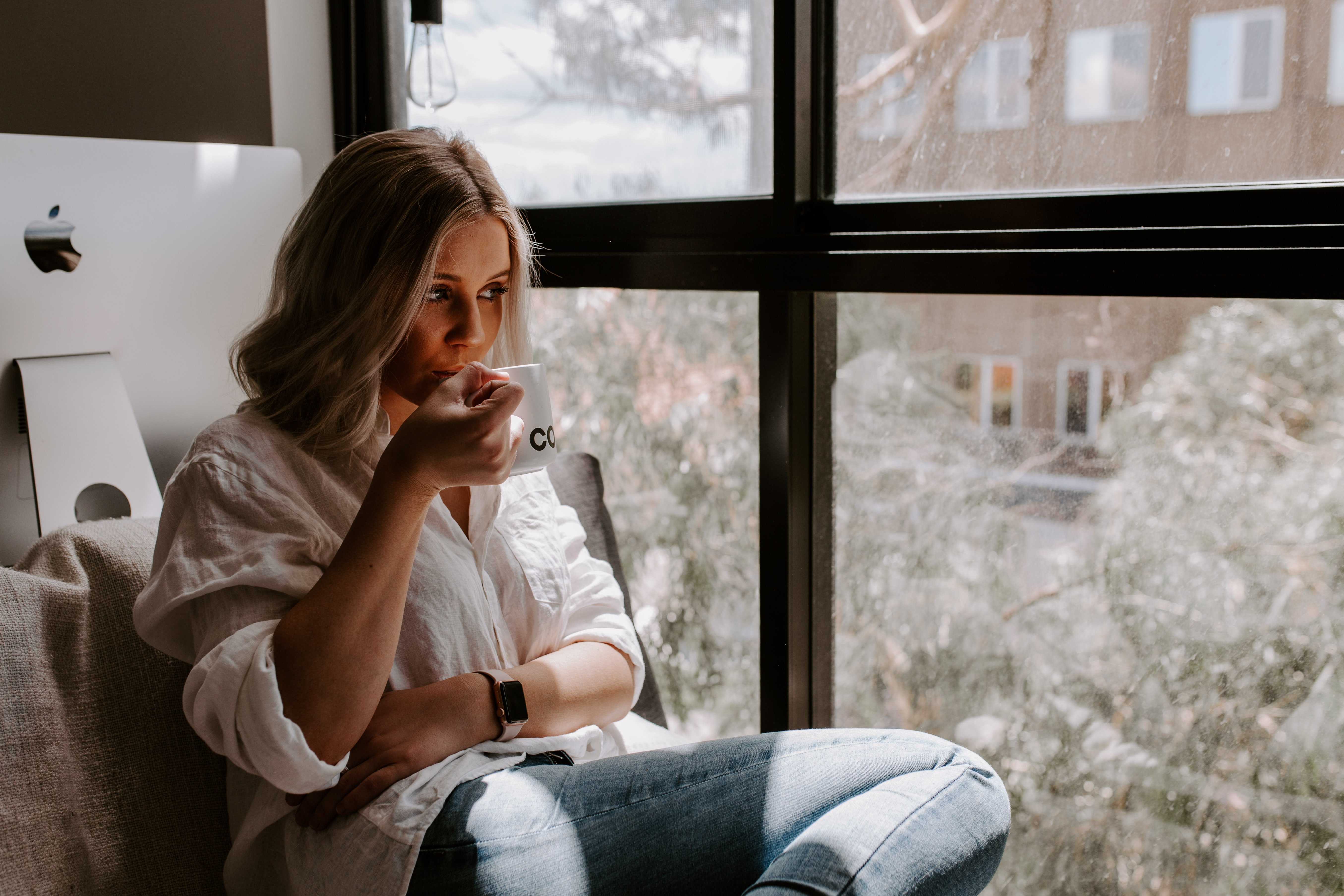 A casually dressed lady sat by a window drinking from a mug