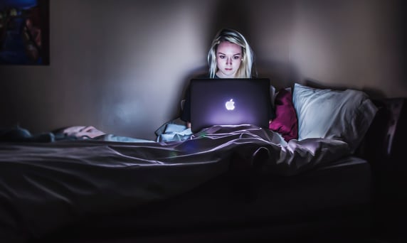 A woman sat on her bed in a dark room looking at her Apple laptop