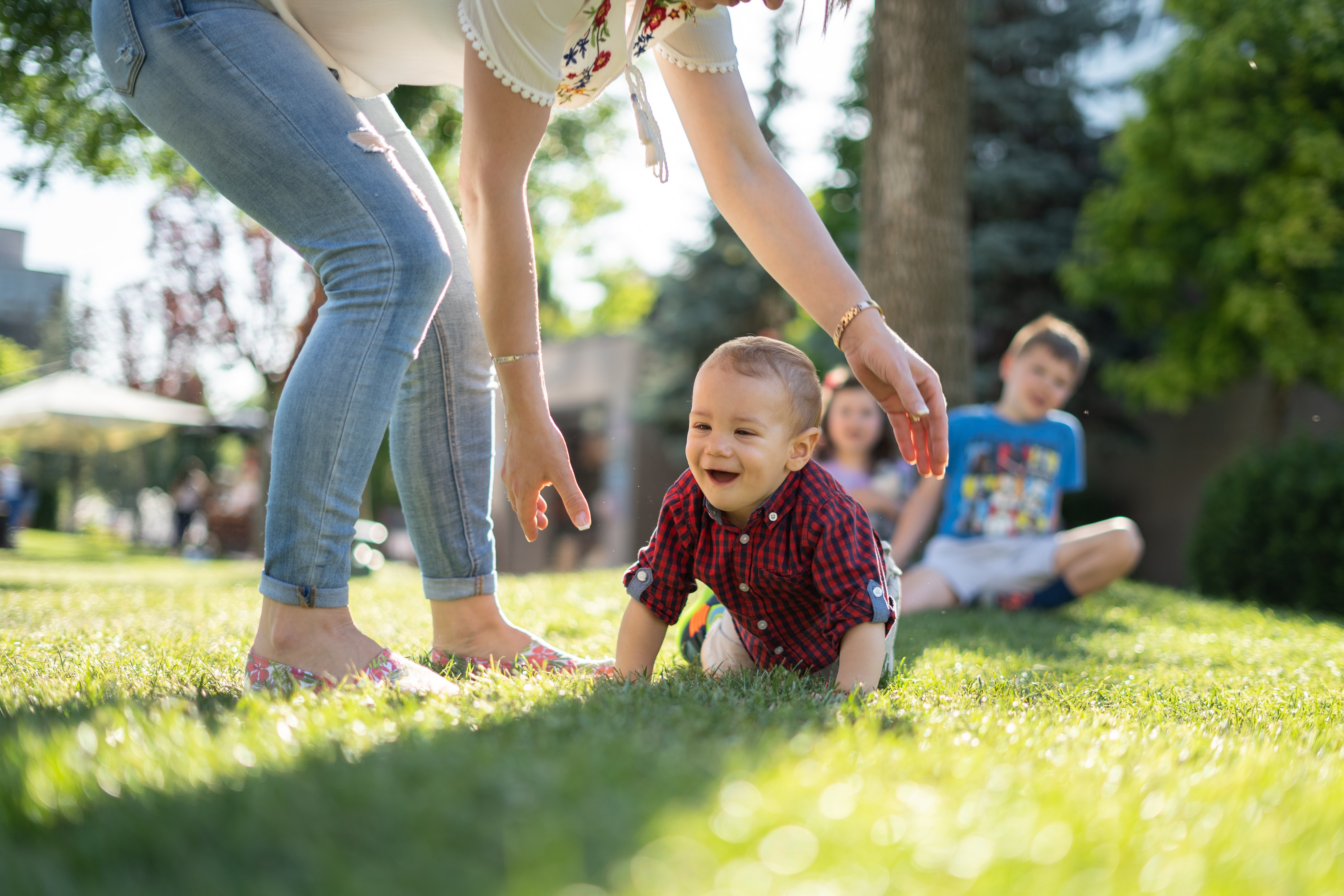 Happy child crawling on the grass towards mum, while his siblings sit behind him