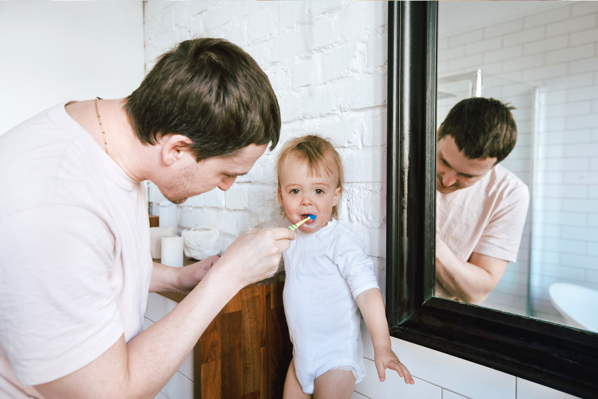 Dad standing in front of a mirror brushing his anxious toddlers teeth