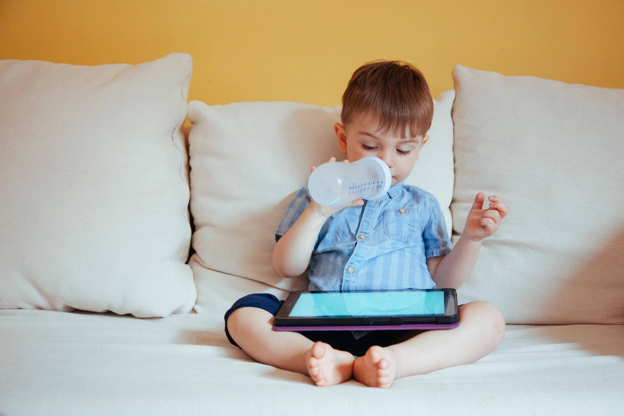 Child sitting on a white sofa looking at an iPad screen while drinking from his bottle