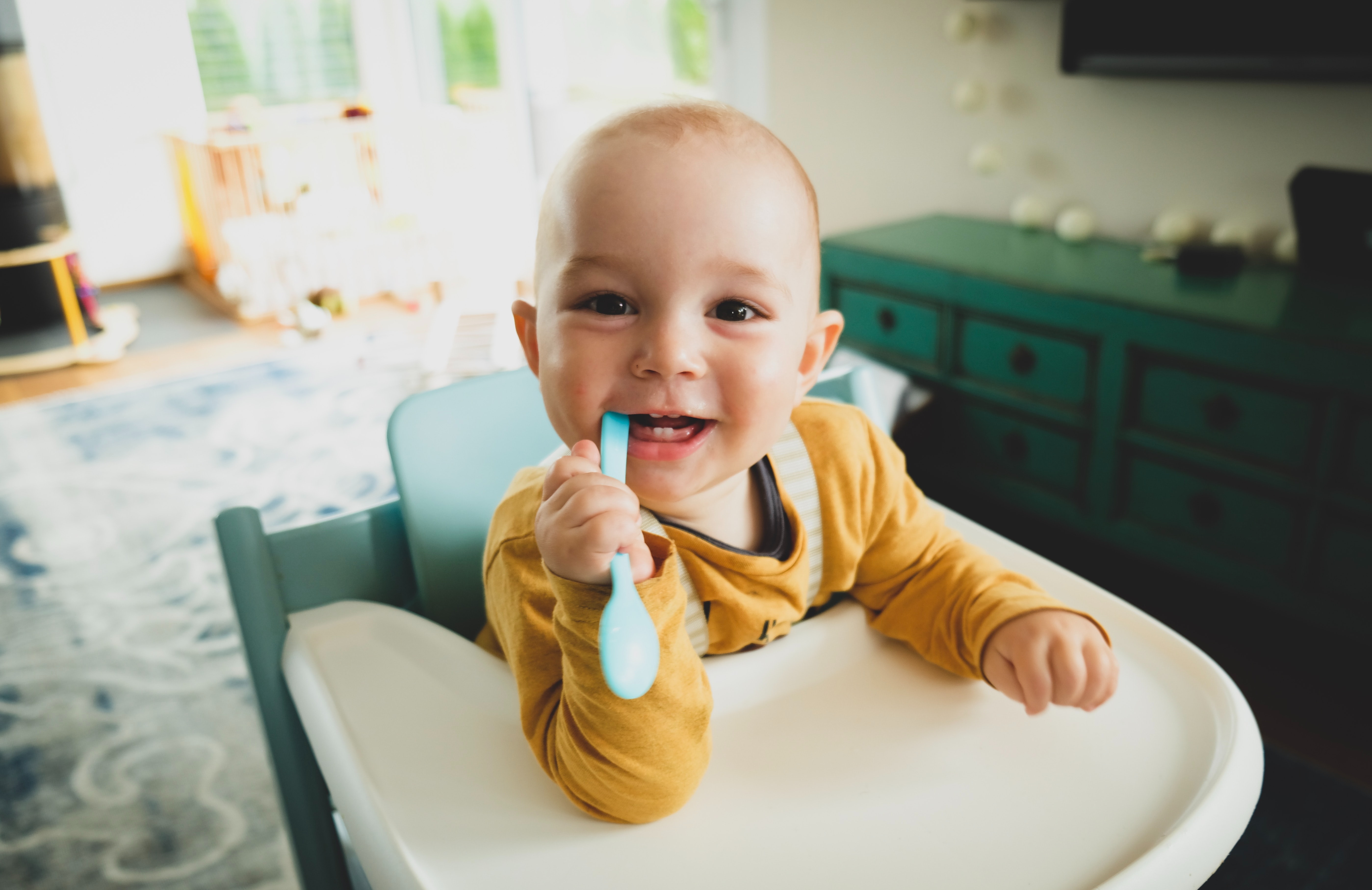 toddler sat in a high-chair smiling and biting on a plastic spoon