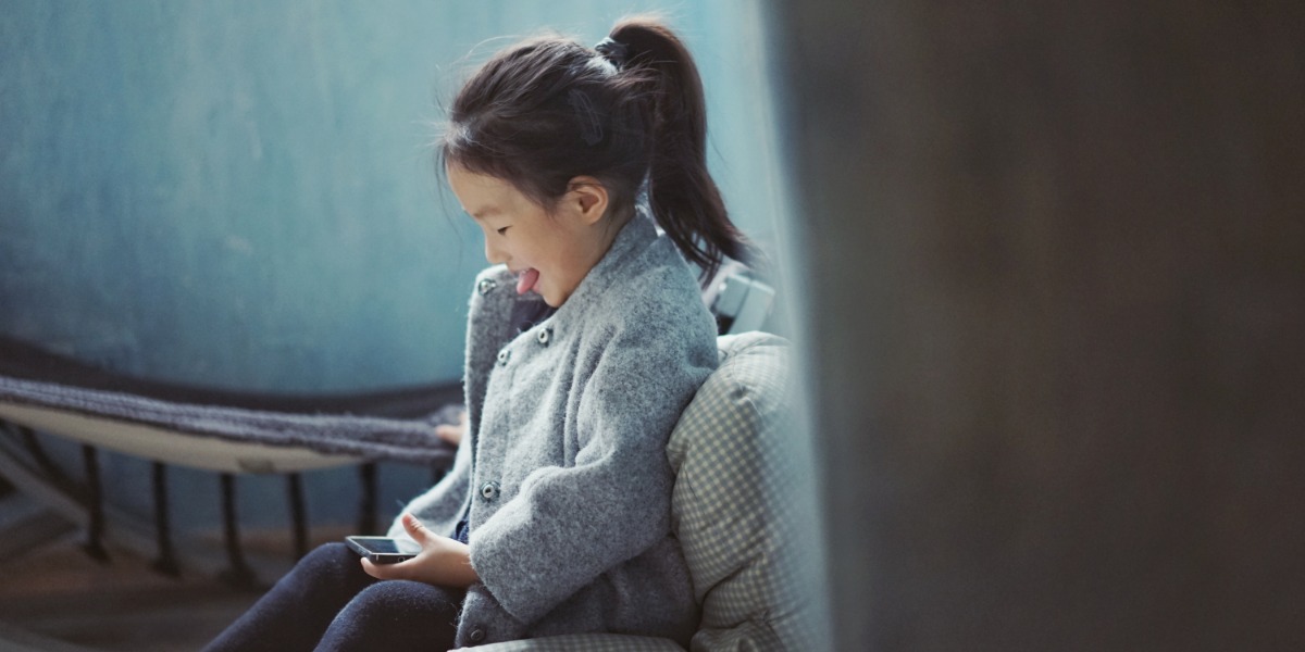 Little girl sat smiling and playing with a device while sat on a seat with a cushion behind her back