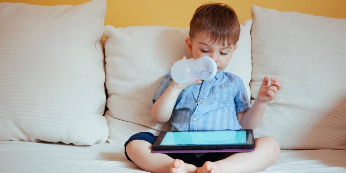 Child sitting on a white sofa looking at an iPad screen while drinking from his bottle