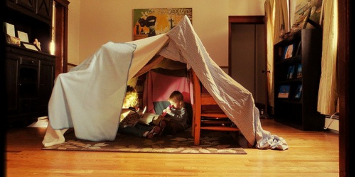 Two children playing in a fort they've built at home using chairs and a sheet