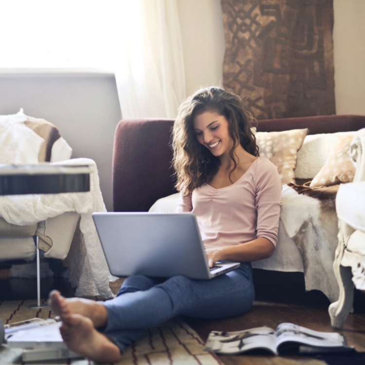 Woman sat on the floor working from home, smiling while using her laptop